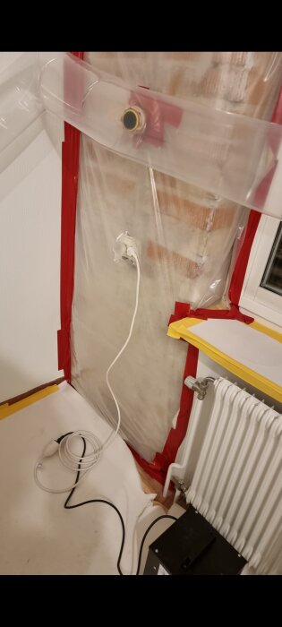 Radonsanering i källare med plastduk, tejp och ventilationsslang. Vitt, röd tejp, fönster, element, kablar syns.