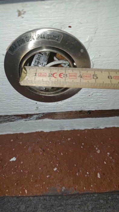 Måttband mäter diameter på rund brandvarnare på vitmålad vägg över brunmålad sockel.
