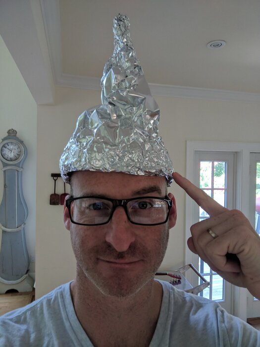 Man med glasögon och hemmagjord hatt av aluminiumfolie inomhus, pekar på hatten, leende, verifieringsförsök.