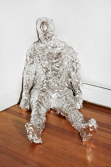 Skulptur i människolik form gjord av aluminiumfolie, sittande mot vit vägg på mörkt trägolv.