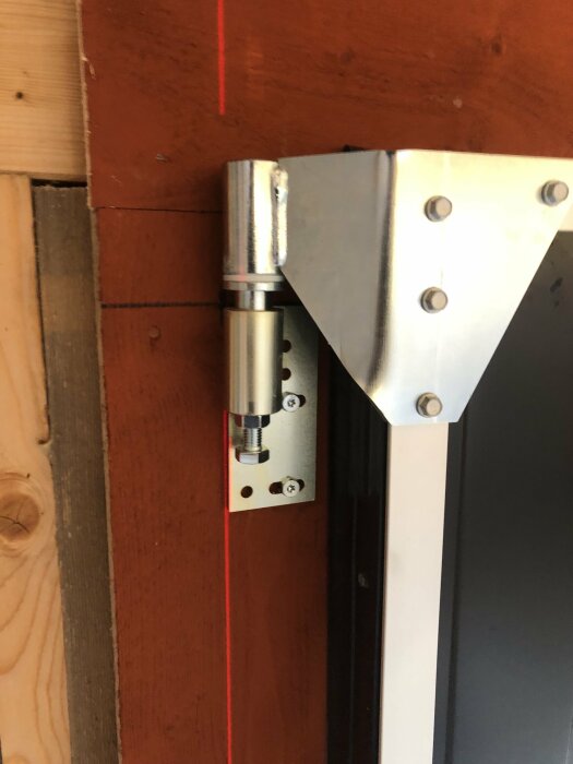 Dörrstängare monterad på röd trävägg och vit dörrkarm, med synlig laserknapp.