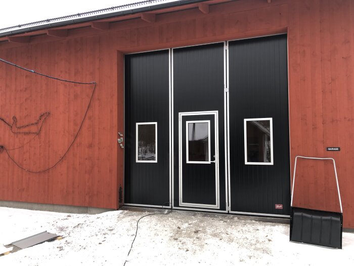 En röd byggnad med svart garageport, vita fönster, snöfläckar på marken och en slang på väggen.