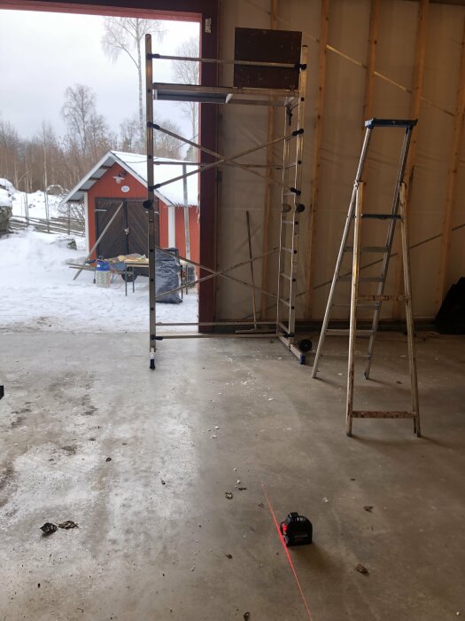Inomhusbyggnadsarbete pågår med ställning, stege och lasernivå; snö utomhus synlig genom öppen port.