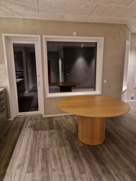 Interiör med träbord, grått golv, vita dörrar och spegelreflektion från fönster. Modernt och minimalistiskt rum.
