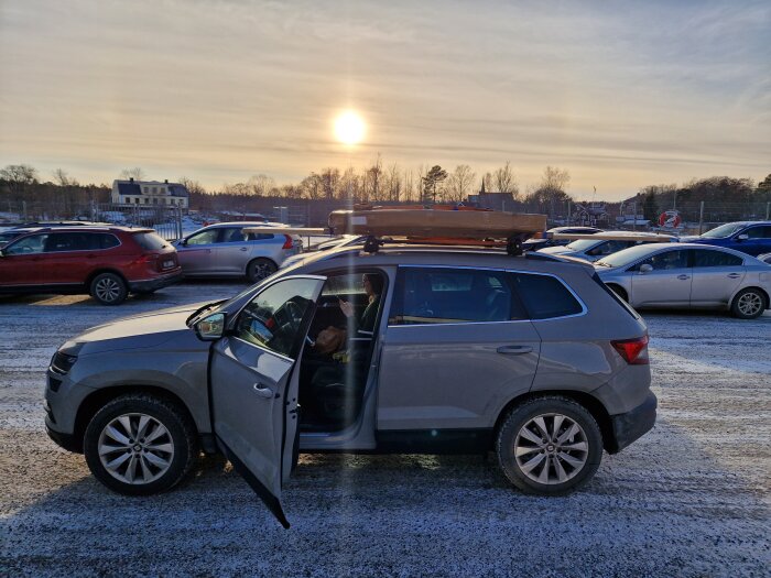 En SUV med öppen dörr, takbox, på en snötäckt parkeringsplats i solnedgång.