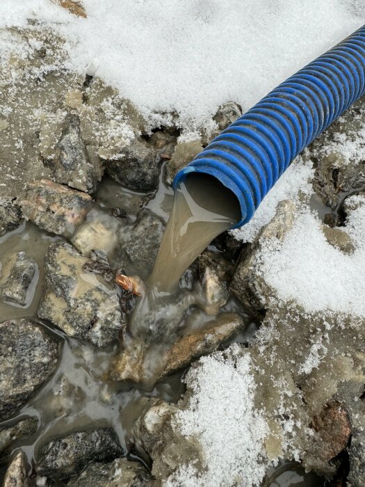 Blå dräneringsslange som tömmer vatten bland smältande snö och stenar.