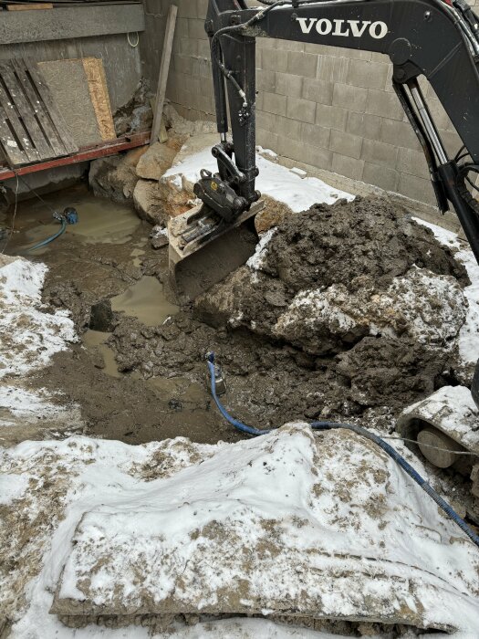 En grävskopa från Volvo gräver i lerig och stenig mark, omgiven av snö och byggmateral, med slaskig vattenansamling.
