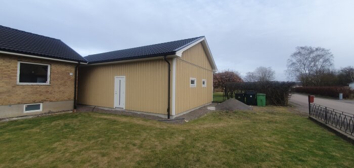 Ett enplanshus med beige panel, svart takpannor, gräsmatta, grusgång, sopkärl och molnig himmel.