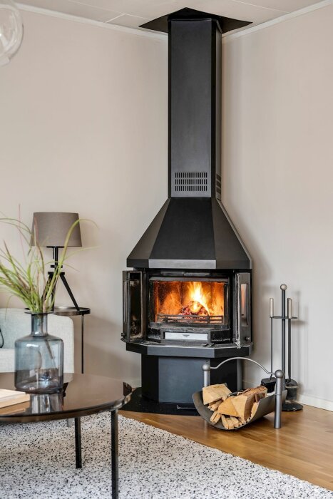 Eldstad med brinnande ved i ett modernt vardagsrum, trägolv, soffa, och bordslampa.