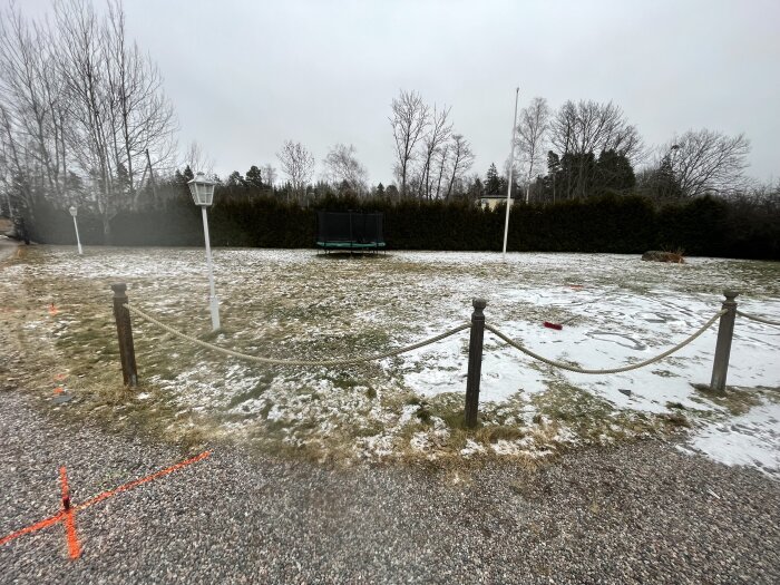 Vinterträdgård med gles snö, gungställning i bakgrunden, repstaket i förgrunden, övergivna leksaker, grå himmel.