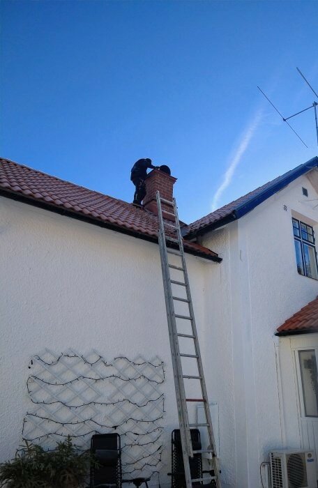 Person på tak arbetar med skorsten, lutande stege mot hus, klart väder.