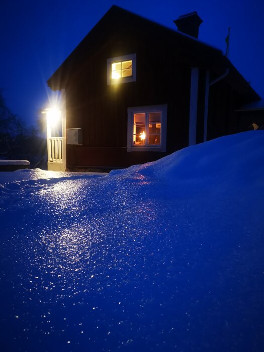 Mysigt trähus med tända lampor och brasa synlig, omgivet av snö i kvällsljus.