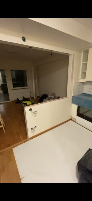 Rum under renovering med öppning mellan kök och vardagsrum, kablar syns, oavslutade väggar, rörigt, kvällsljus.