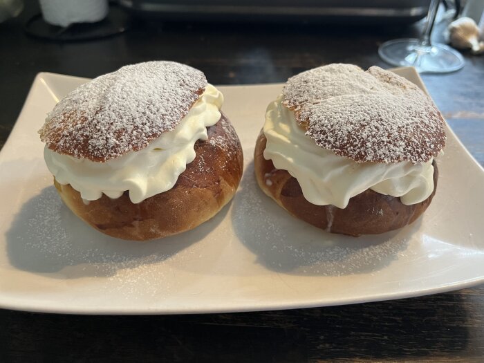 Två semlor med vispgrädde och florsocker på en vit tallrik.