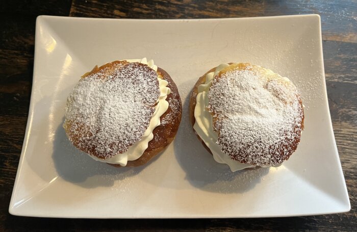 Två semlor med florsocker på vit tallrik och mörkt bord.