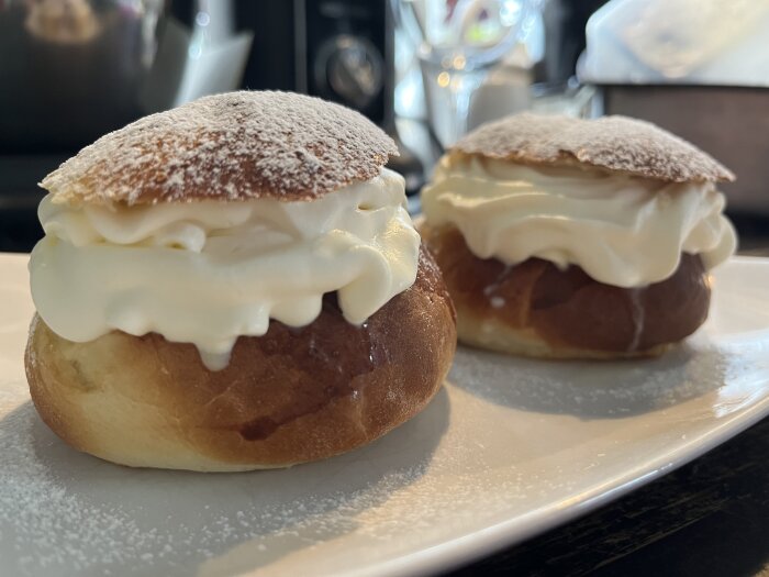 Två semlor med fluffig grädde och pudrat florsocker på en vit tallrik.