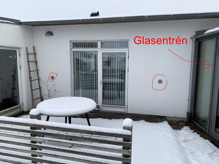 Terrass med snö, rundat bord, stegar, skjutdörr med text "Glasentrén", vägglampor. Vinter eller kallt klimat.