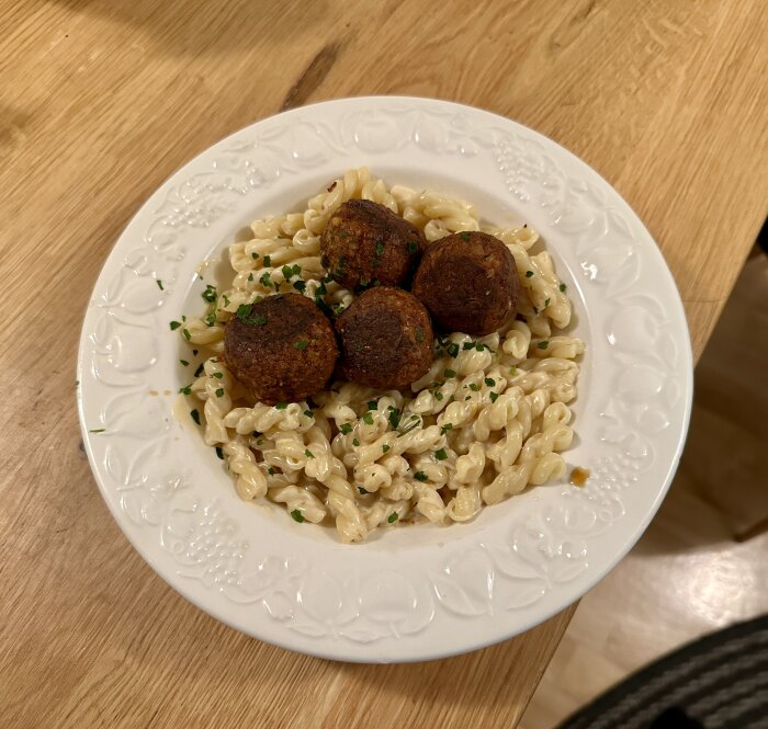 Pasta med köttbullar och hackad persilja på vitt mönstrat tallrik på träbord.