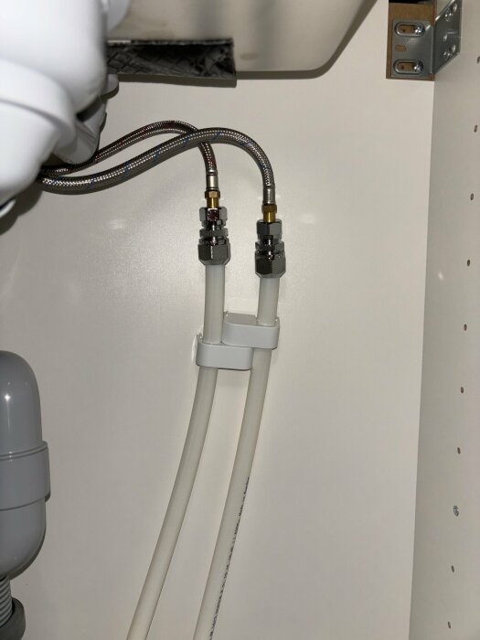 VVS-installation med flexibla slangar och kopplingar under diskbänk, grått avloppsrör, vit skåpvägg.