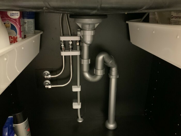 Ett underskåp med vaskens VVS-installationer, avloppsrör, vattenlås och anslutningar i hemmiljö.