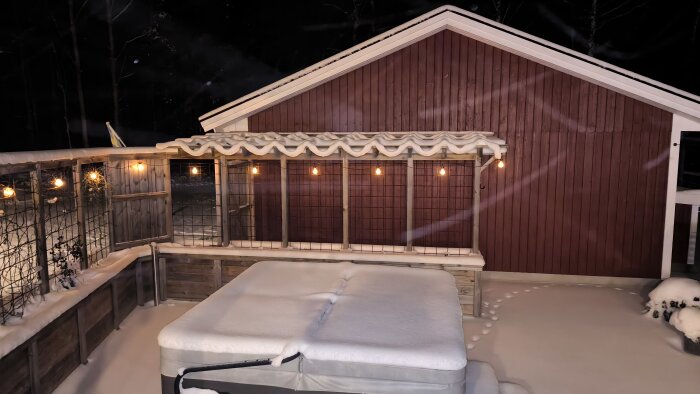 Snötäckt utomhusspa, belyst veranda och röd stuga på natten i vinterscenario.