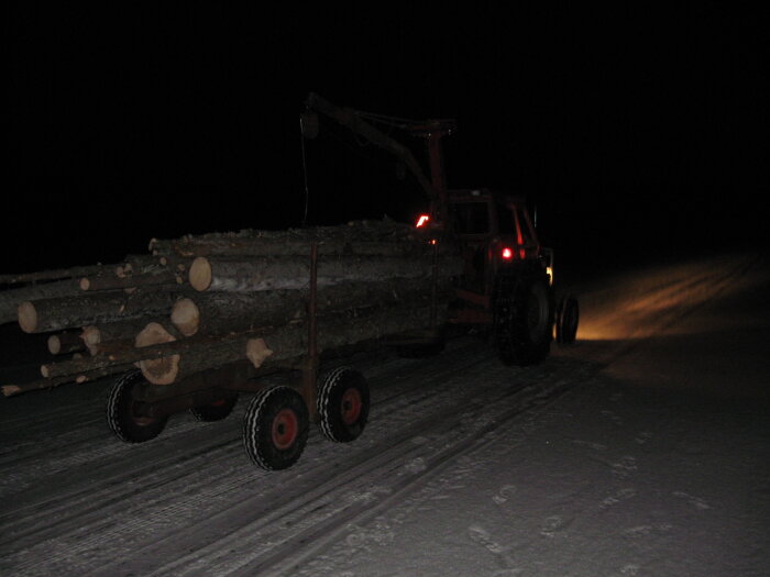 Lastbil med timmer kör på snöig väg nattetid, med släp och baklyktor som lyser.