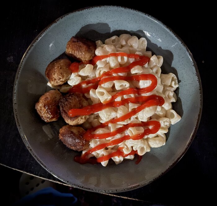 En tallrik med macaroni and cheese och köttbullar, toppad med ketchup, serveras på ett mörkt bord.