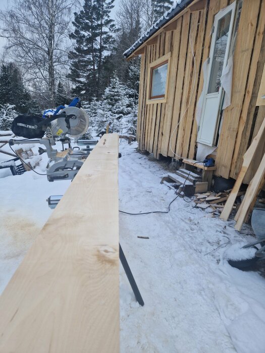 Snöig arbetsplats utomhus med såg och träplanka framför timmerbyggnad, i skogsmiljö.