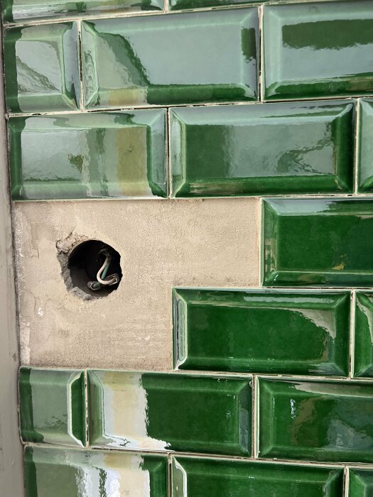 Gröna kakelplattor på en vägg med ett hål där elledningar är synliga.
