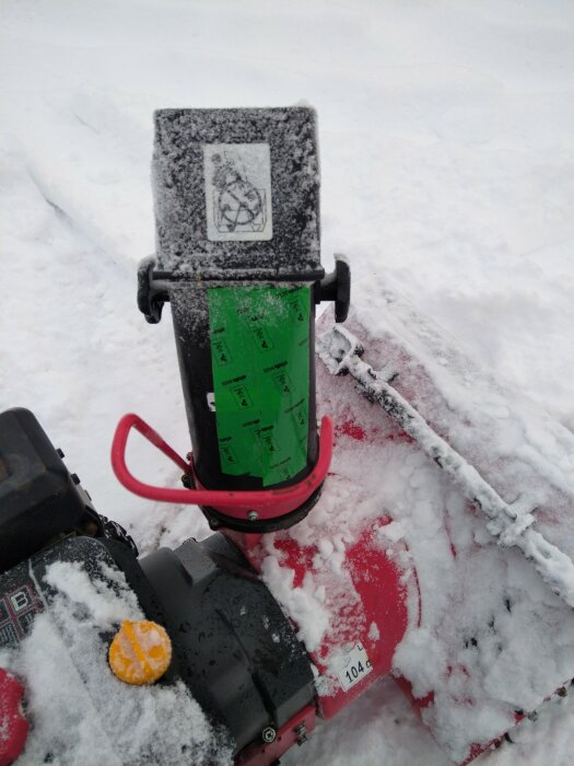 Snöig bensindriven snöslunga med varningsdekal, röda detaljer och synligt kontrollnummer, parkerad utomhus.