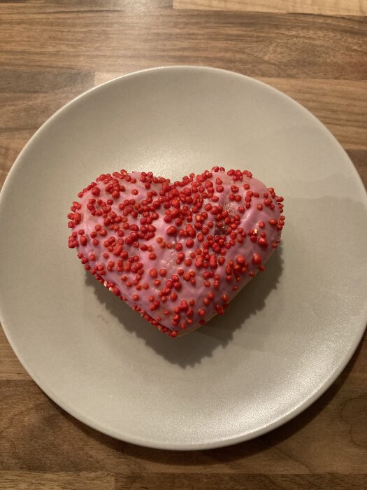 Hjärtformat bakverk med rosa glasyr och röda strössel på en grå tallrik.