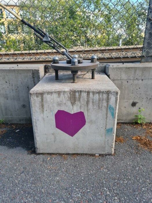 Betongfundament med metallkonstruktion, staket bakom, lila hjärtmålning, stadsmiljö, mark, vegetation, ingen person synlig.