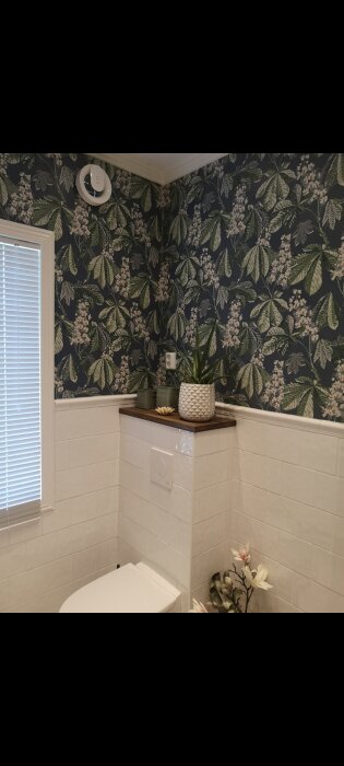 Ett modernt badrum med grönt växttryck på tapeten och vita kaklade väggar.