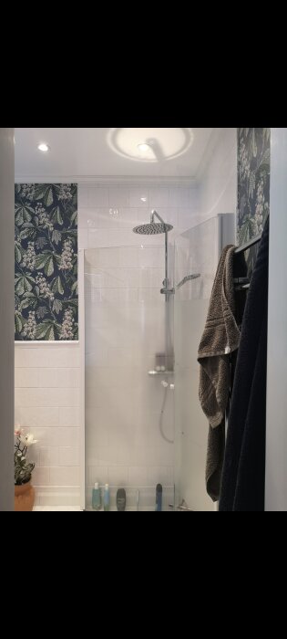 Modernt badrum med glasduschutrymme, vita kakelväggar, mörkblå tapet med växtmotiv och upphängda handdukar.