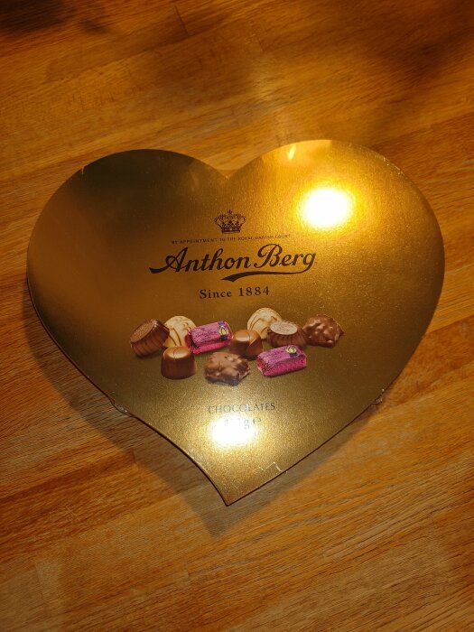 Hjärtformad Anthon Berg chokladask på träbord. Guldig med bilder av praliner. "Since 1884".