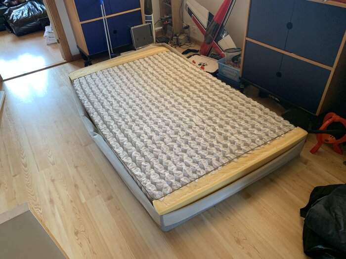 En säng utan lakan i ett rörigt rum med möbler och elektronik syns.