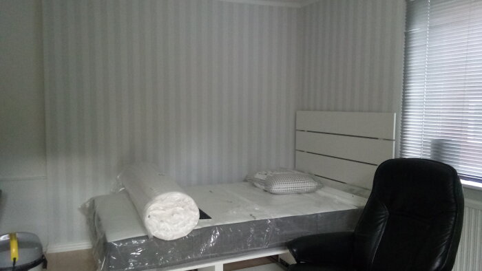 Ett minimalistiskt och neutralt sovrum med säng, byrå och kontorsstol, med randig tapet och stängda persienner.