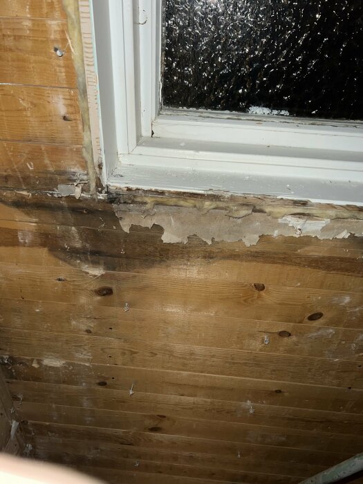 Fönster med fuktskador och avskalad färg på träpanelen inne, natt och regndroppar syns ute.