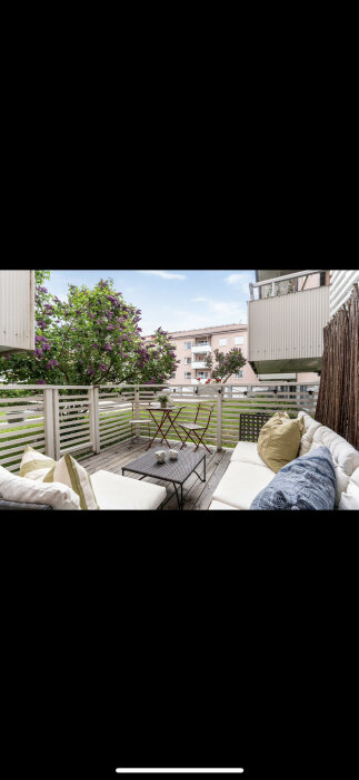 Ett mysigt balkongutrymme med soffa, kuddar, bord, lila blommor och utsikt över bostadsbyggnader.