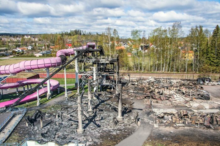 Förstörd byggnad vid vattenpark, rosa rutschkanor, brända rester, urbant landskap i bakgrunden.