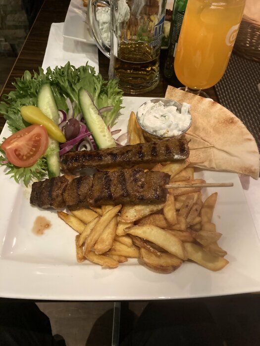 En tallrik med souvlaki, pommes frites, sallad, tzatziki och pita. Öl och apelsinjuice i bakgrunden.