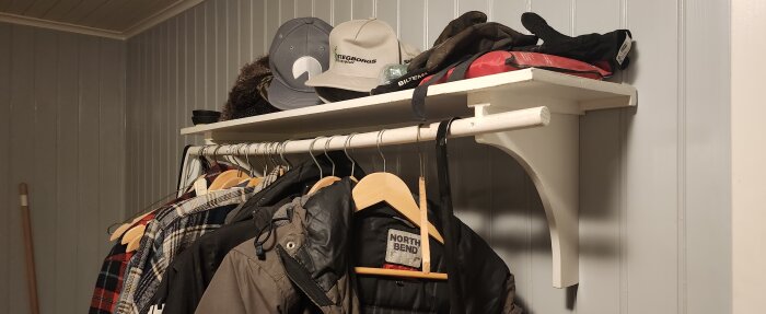 En hallhylla med kläder på galgar, hattar och handskar, grå väggar i bakgrunden.