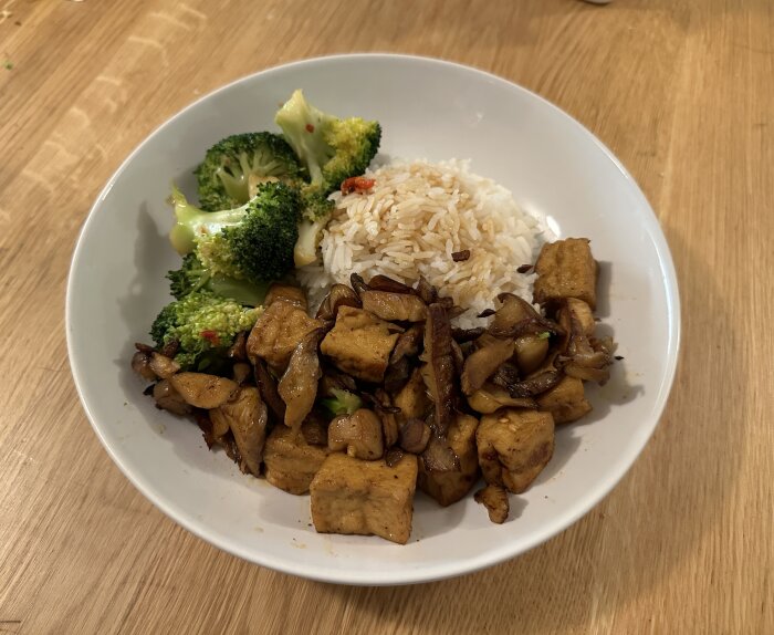 Vit tallrik med ris, stekt tofu, svamp och ångad broccoli, serverat på träbord.