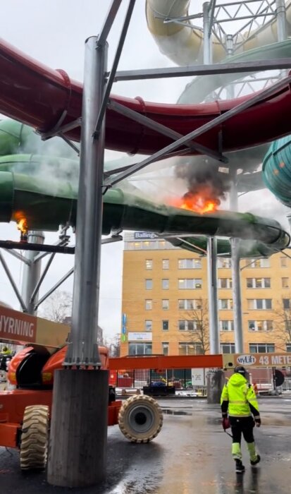 Vattenrutschkana på brand, räddningspersonal på plats, konstruktionsområde, rök och eldslågor synliga, stadsmiljö.