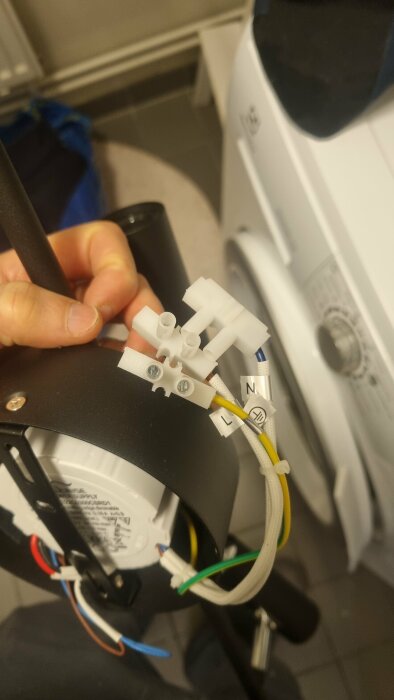 Person håller en elektrisk enhet med oanslutna kablar, tvättmaskin i bakgrunden, installation eller reparation pågår.