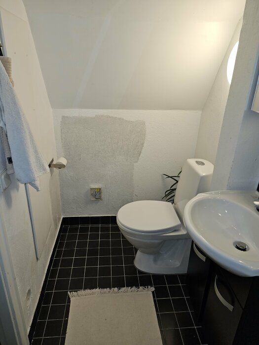 Badrum med toalett, handfat, matta och handdukar. Vägg reparationsområde synligt. Svartvitt golv, inramat av vita väggar.
