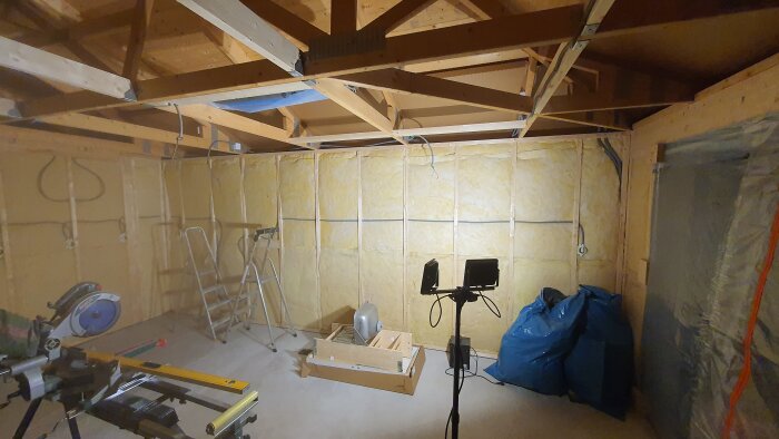 Byggarbetsplats inomhus med synlig isolering, trästomme, verktyg och belysningsutrustning.