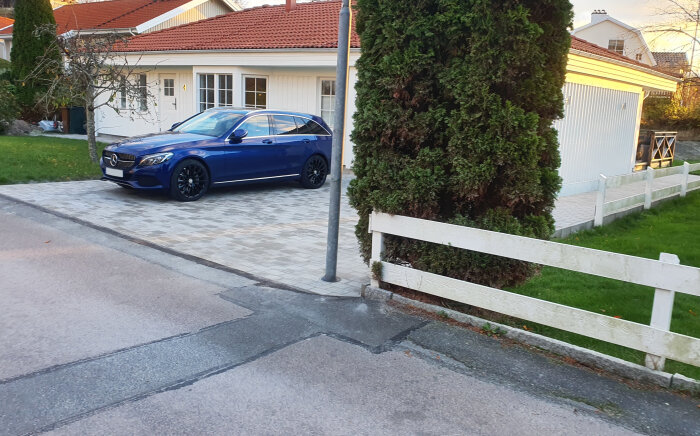Blå bil parkerad framför hus, vit staket, garage, stenlagd uppfart, träd, gräsmatta, gatlyktpåle, krukor.