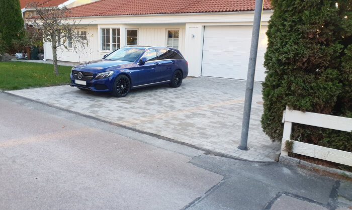 En blå bil parkerad framför ett vitt garage intill en häck vid ett hus.