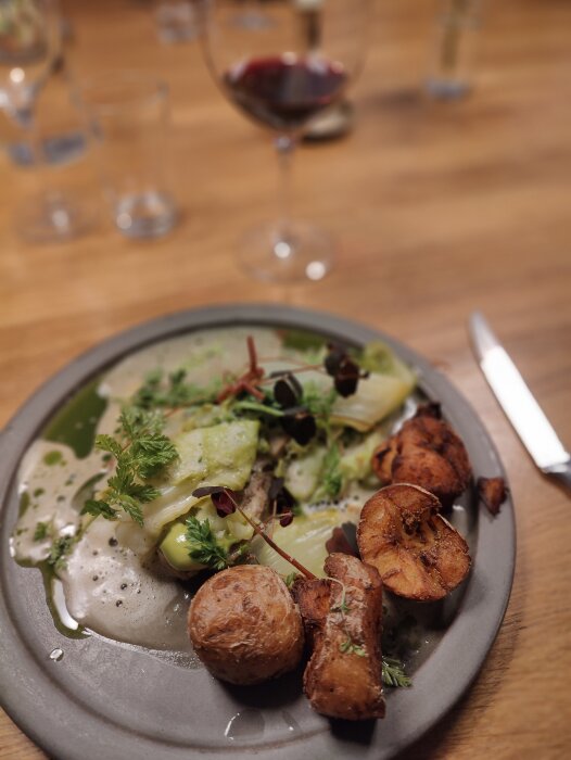 Middagstallrik med grönsaker, bakad potatis, svamp och sås. Vin i bakgrunden. Oskärpa skapar intimitet. Elegant presentation.